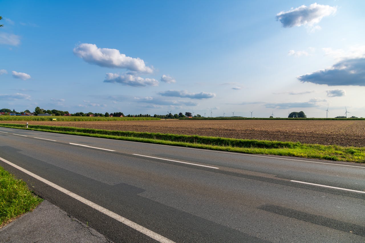A sunny rural scene with an empty road, open fields, and wind turbines in the distance.