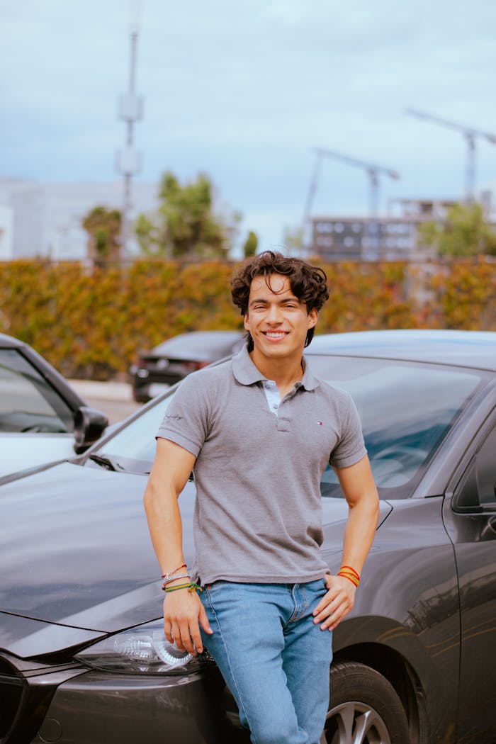 Young man in casual attire leans on a car, smiling in a parking lot setting.