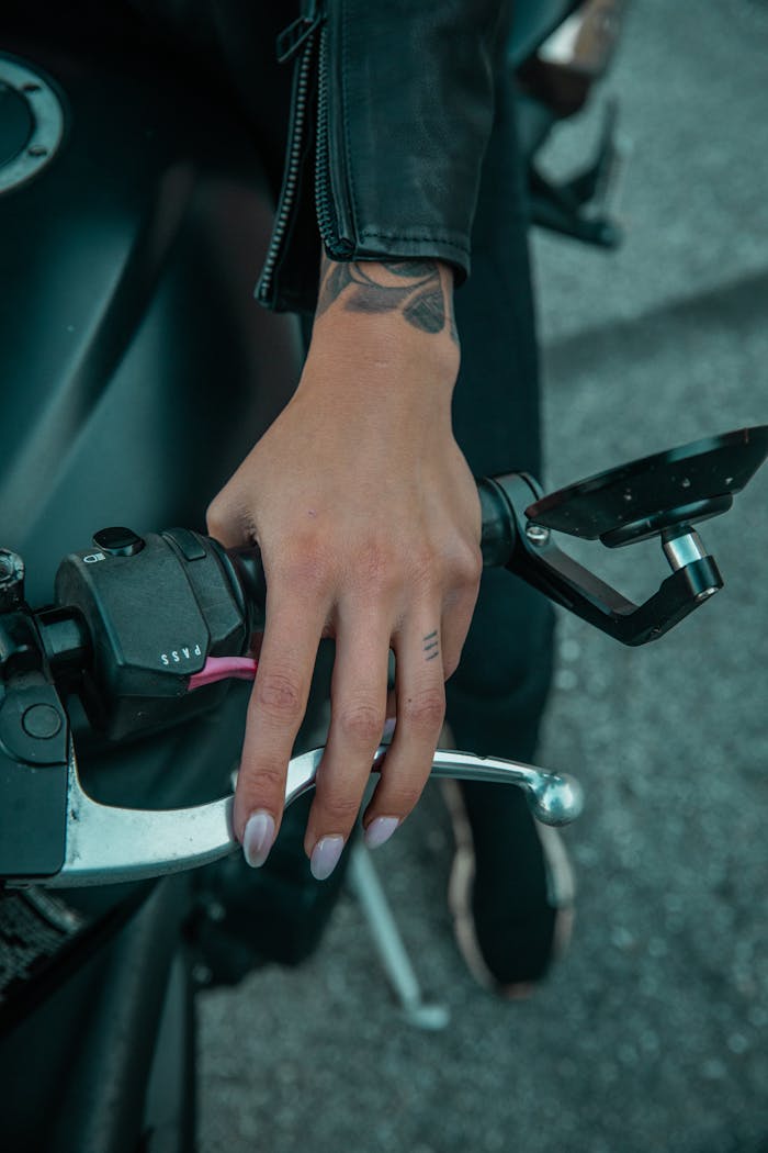 A detailed view of a hand gripping a motorcycle handlebar, showcasing tattoos and leather jacket.